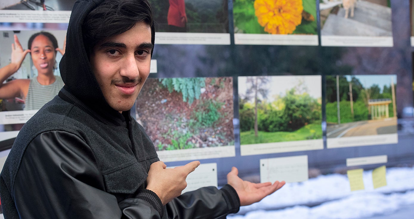 Hamza in a dark jacket with the hood up on his head. He's pointing slightly to his left at the photos on display