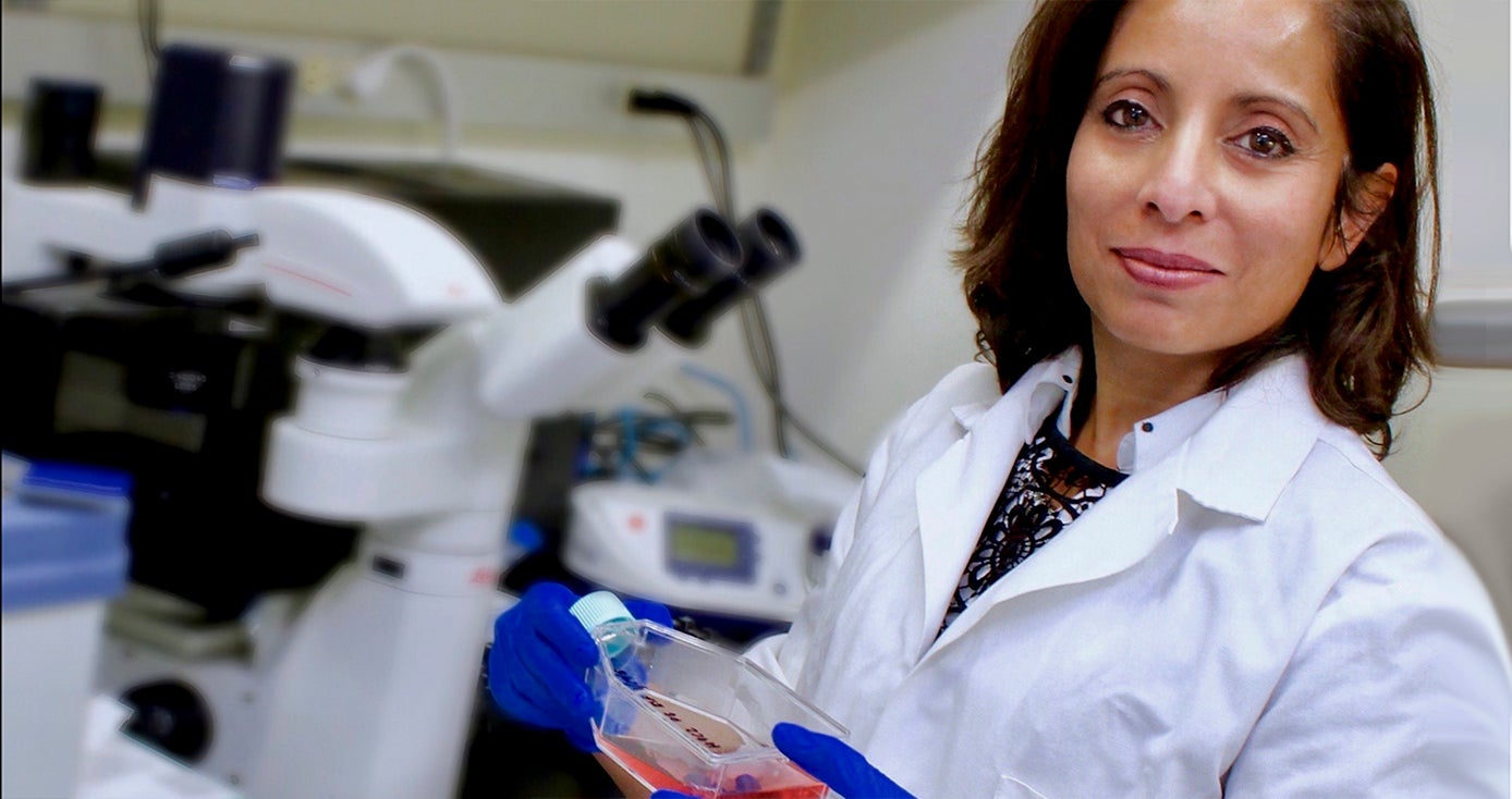 Maliha Zahid in white lab coat and wearing blue gloves holding a clear bottle of liquid in a lab setting