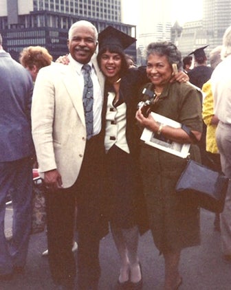 a woman in graduation garb with her arms over her parents' shoulders: dad in a white suit, mom in a green dress