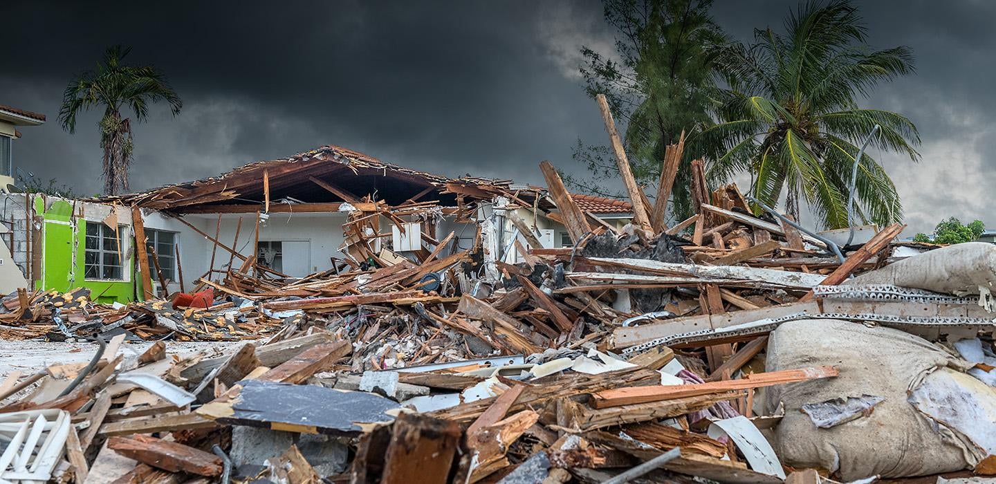 A house is destroyed after a hurricane. The sky is dark and cloudy, and palm trees blow in the wind