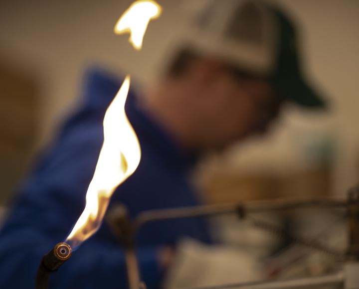 A blowtorch releases flame, with Ryan Tate in background