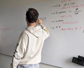 A student in a beige hoodie writes on a dry erase board