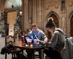 Two students study at a table in the Cathedral of Learning Commons Rooms. Two Christmas trees stand by the entrance.