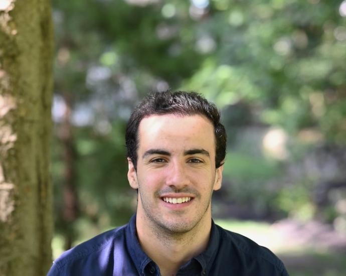 Man with dark hair sits in a wooded area and smiles for camera.