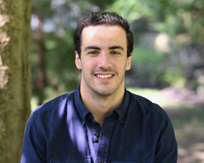 Man with dark hair sits in a wooded area and smiles for camera.