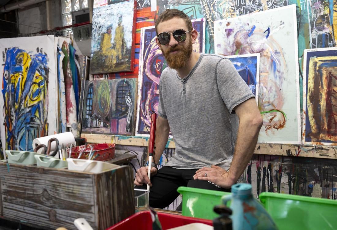A man wearing sunglasses, surrounded by abstract paintings.