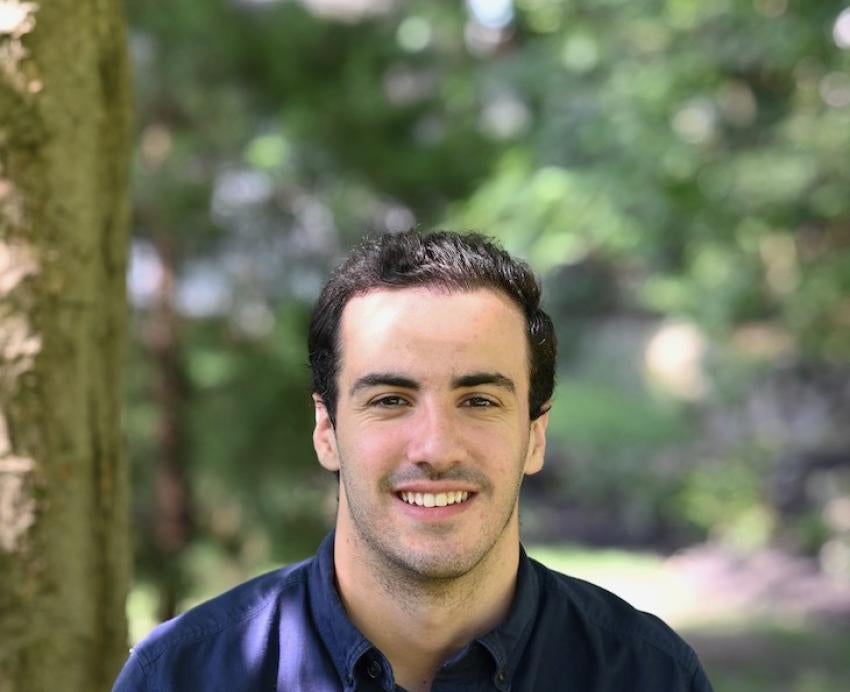 Man with dark hair sits in a wooded area and smiles for camera.